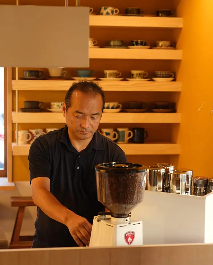 カフェオーナーの男性がコーヒー豆グラインダーを操作する、温かみのあるカフェのカウンター風景